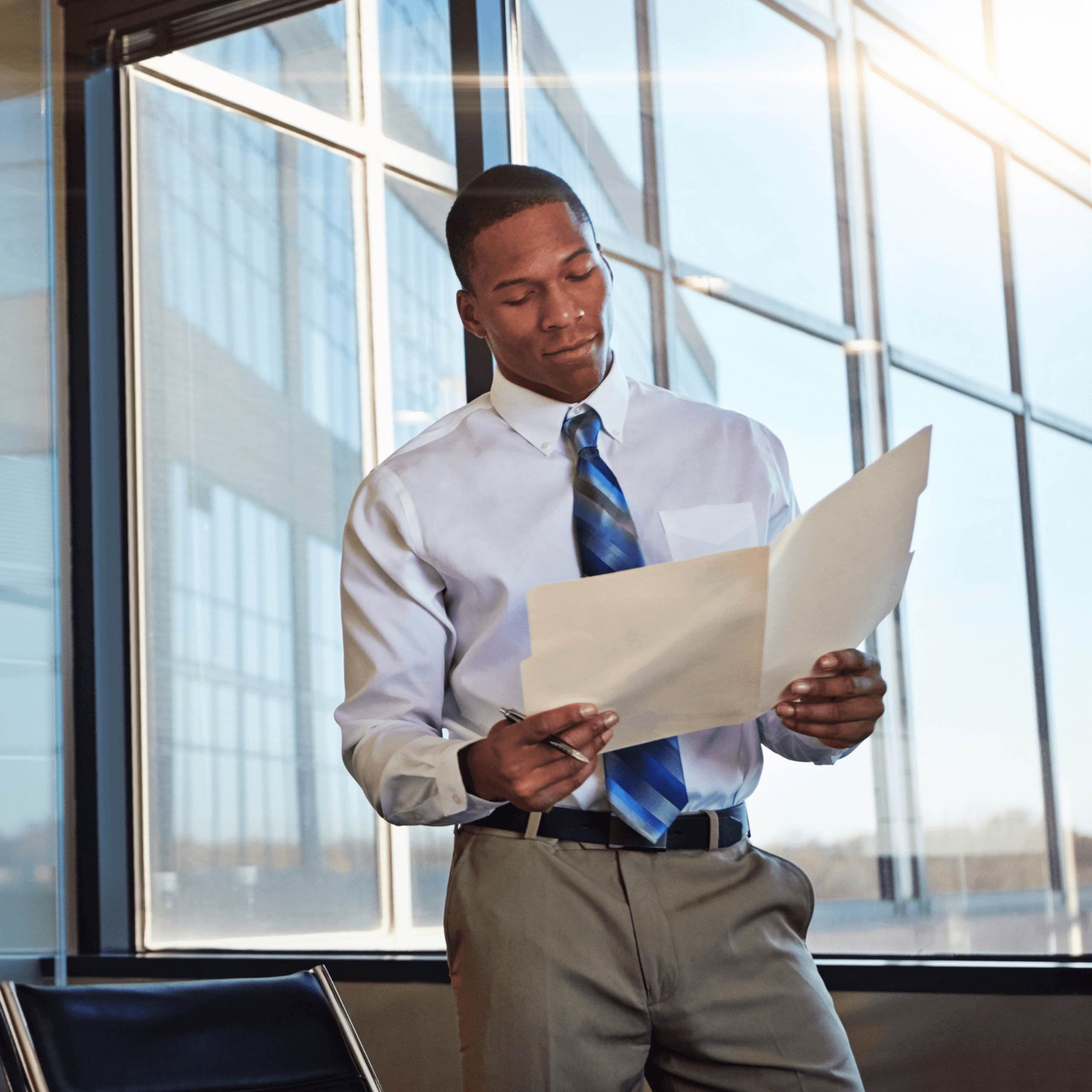 A man in business attire stands by large office windows, holding and reviewing documents while the sun shines outside.