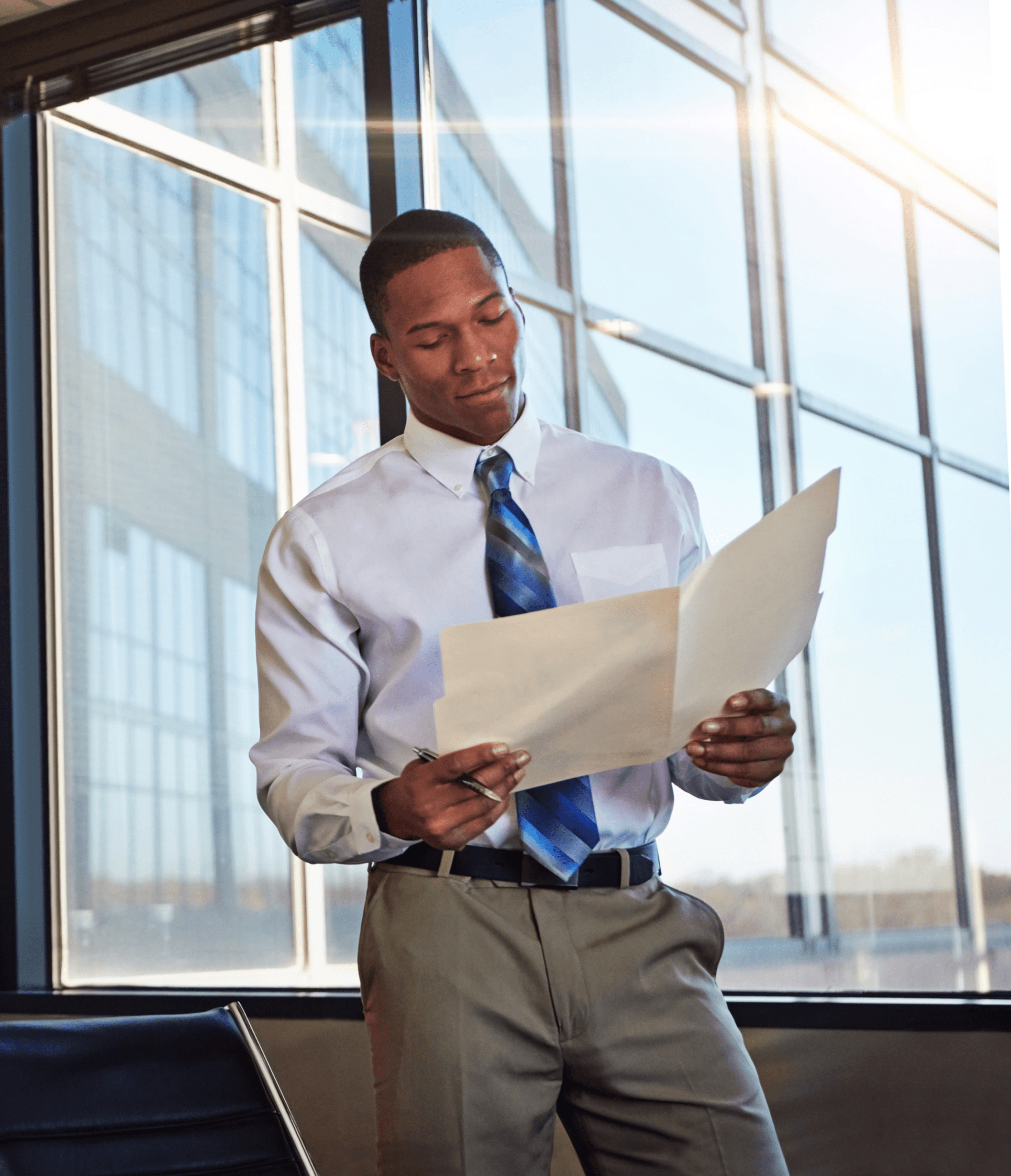 A man in business attire stands by a large window, reading documents. Sunlight streams in, creating reflections on the glass.