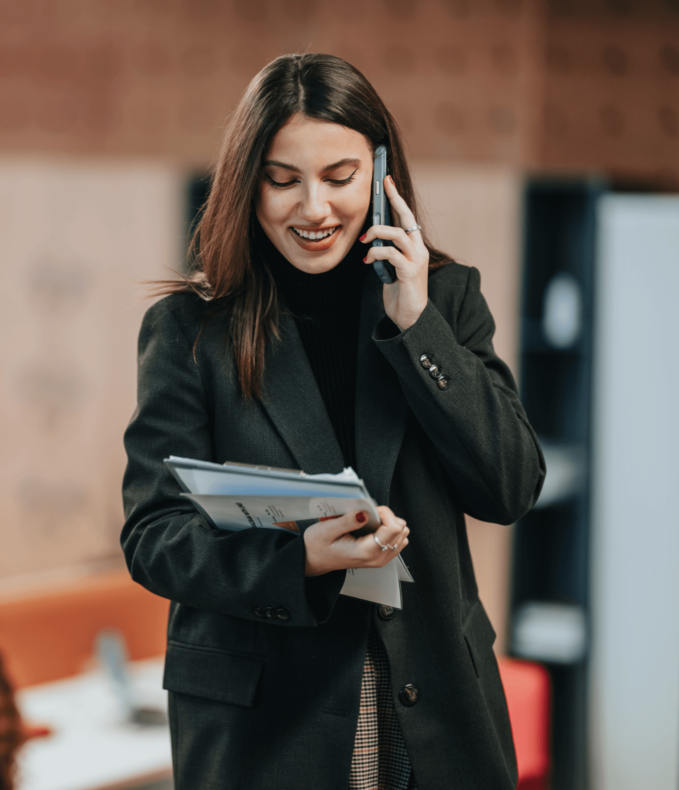 A woman wearing a dark blazer is smiling while talking on her smartphone and holding several documents in an office setting.