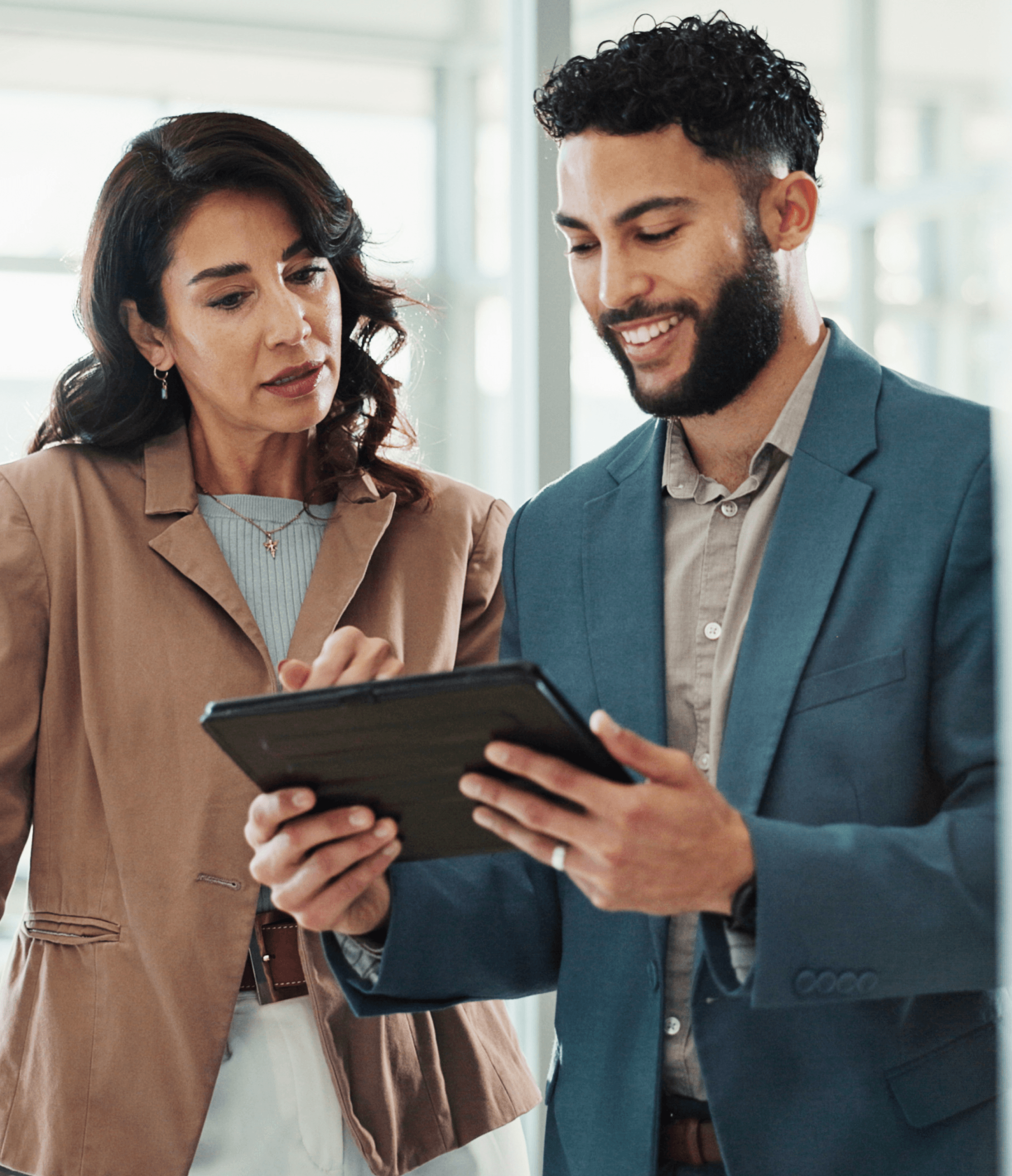 Two professionals stand together, looking at a tablet and discussing something. The woman wears a tan blazer, while the man wears a blue blazer. They appear focused and engaged in a modern office setting.