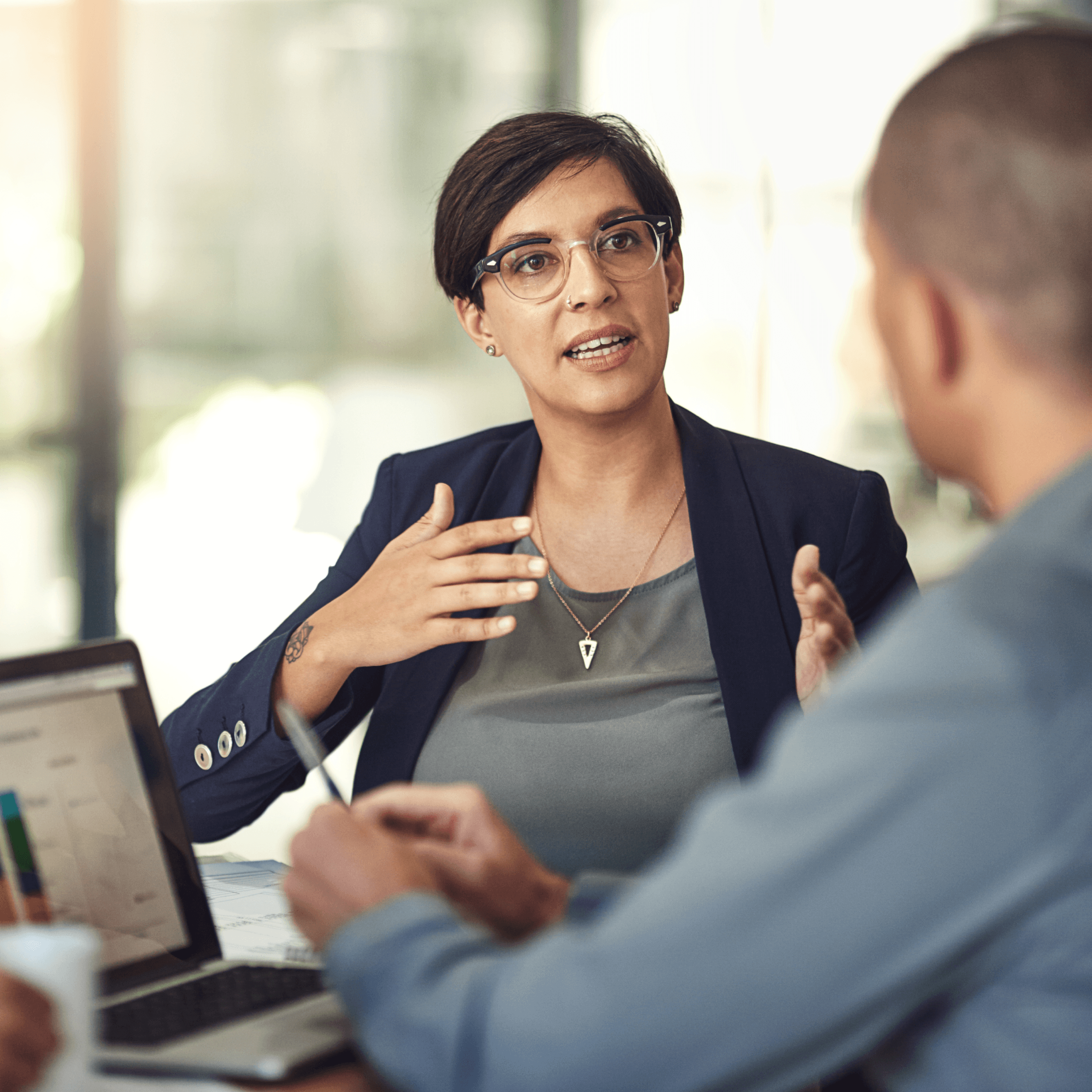A woman wearing glasses and a blazer gestures while talking to a man during a meeting. A laptop and documents are on the table, suggesting a professional office setting.